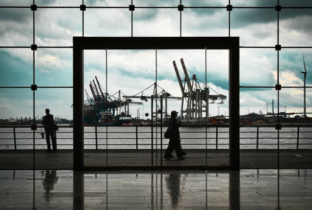 View through a large window front onto pedestrians as the imposing cranes of the Hamburg harbor reflect the vibrant atmosphere of the port city in the background.