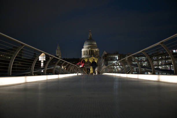 The iconic dome of St. Paul’s Cathedral spotlit at night overlooking the futuristic glass and steel span of the Millennium Bridge crossing the River Thames in the heart of London, UK.