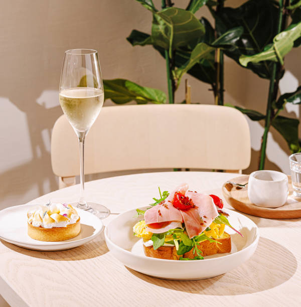 A brunch setting features a plate of colorful ingredients topped with sliced meats and greens, accompanied by a dessert and a glass of sparkling beverage. It's a cozy atmosphere.