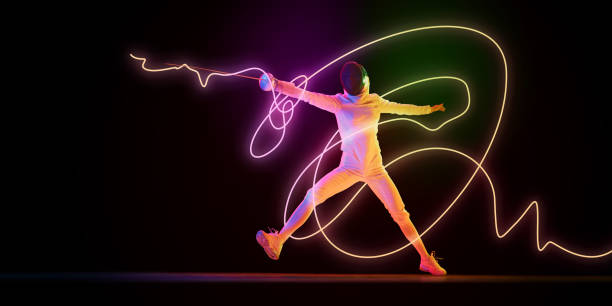 istock Fencer in white uniform lunging with sword extended, neon orange and green light streaks tracing movement against dark background. 2200397577