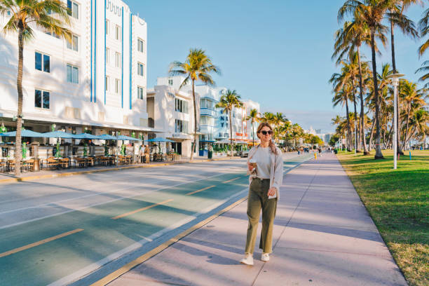 Woman holding coffee walks along Ocean Drive in Miami Beach, Art deco Historic district Florida USA A woman in casual attire walks along Ocean Drive in Miami Beach, holding a coffee, with iconic Art Deco buildings, palm trees, and outdoor cafés creating a sunny, relaxed atmosphere. 1 hotel south beach stock pictures, royalty-free photos & images