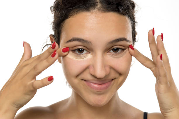 istock Young woman with concealer under eyes, smiling. Happy expression during beauty routine. Close-up studio portrait highlighting skincare application on white background 2200417392