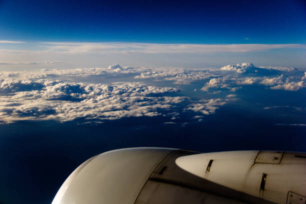 A stunning aerial view from an airplane window shows a vast expanse of fluffy cumulus clouds stretching to the horizon beneath a deep blue sky. The upper atmosphere is a clear, brilliant blue, gradually transitioning to a lighter shade where it meets the cloud layer. The clouds themselves are a mix of bright white and darker grey, suggesting varying thicknesses and levels of sunlight penetration. Part of the airplane's engine cowling is visible in the lower right corner, providing a sense of scale and perspective. The overall mood is serene and majestic.