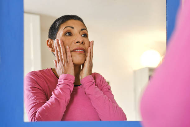 Beautiful non-binary person touching their face while looking in the mirror at home stock photo