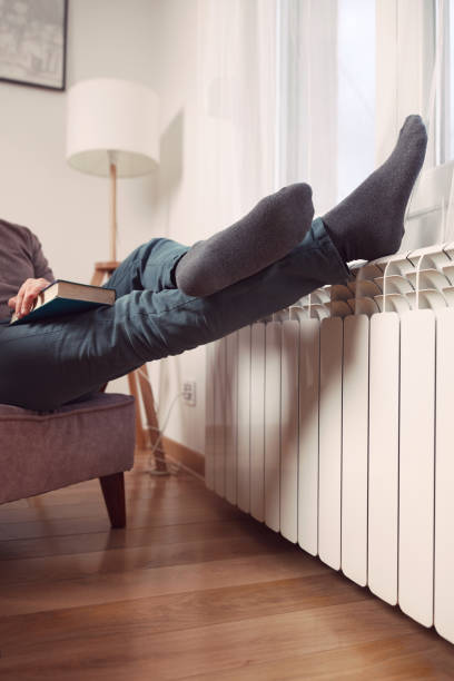 istock Man's feet with socks on a home heating radiator on a cold winter day outdoors while reading a book. 2200427024