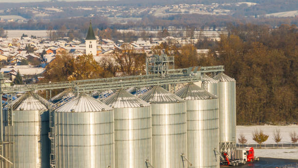 A large, metallic silo stands prominently in a snow-dusted agricultural field, surrounded by a picturesque winter village with a distant forest and hills under a clear sky.