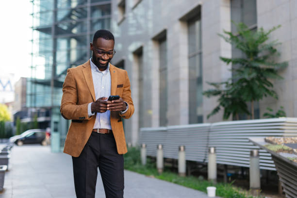 istock Young businessman in brown blazer checking smartphone outside office building 2200428633