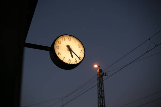 A glowing station clock shows the time against the deep blue evening sky, with power lines and a lit streetlamp in the background. The contrast between artificial light and the fading natural light creates a moody and atmospheric urban scene. The clock serves as a focal point, symbolizing time, travel, and anticipation.