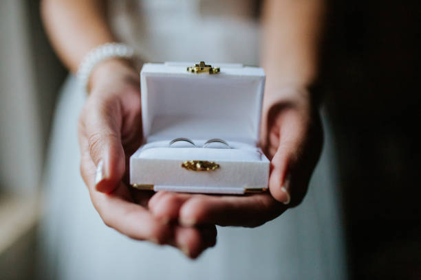 A bride gently holds an open white box with two wedding rings, symbolizing love and commitment. The soft lighting and delicate details capture the intimate beauty of the moment.
