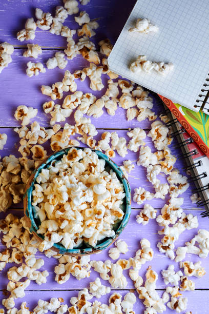 Top view on clay bowl with popcorn on purple workspace (desk) with pencil and notebook