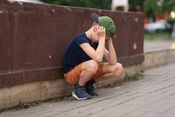 A teenage boy is squatting leaning against a concrete wall on the street. Upset and sad. Bullying. Quarrel with parents A teenage boy is squatting leaning against a concrete wall on the street. Upset and sad. Bullying. Quarrel with parents. sad teenager boy crying outdoors in park stock pictures, royalty-free photos & images