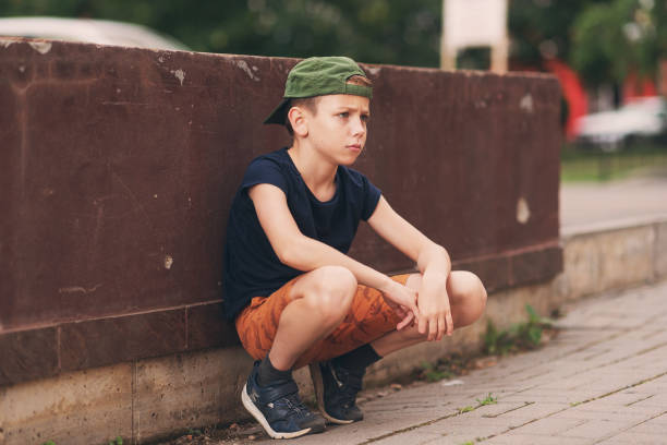 A teenage boy is squatting leaning against a concrete wall on the street. Upset and sad. Bullying. Quarrel with parents A teenage boy is squatting leaning against a concrete wall on the street. Upset and sad. Bullying. Quarrel with parents. sad teenager boy crying outdoors in park stock pictures, royalty-free photos & images