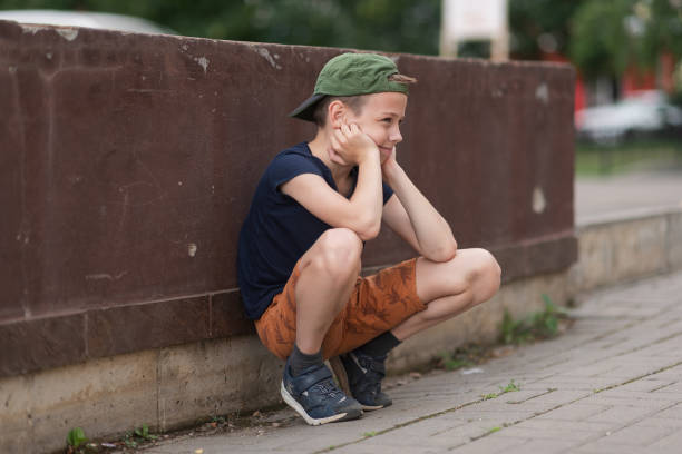 A teenage boy is squatting leaning against a concrete wall on the street. Upset and sad. Bullying. Quarrel with parents A teenage boy is squatting leaning against a concrete wall on the street. Upset and sad. Bullying. Quarrel with parents. sad teenager boy crying outdoors in park stock pictures, royalty-free photos & images