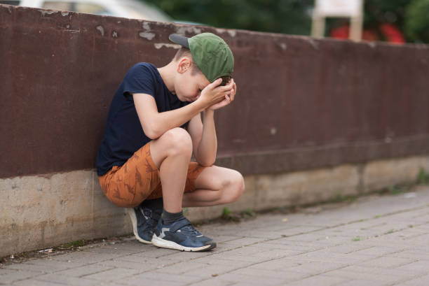 A teenage boy is squatting leaning against a concrete wall on the street. Upset and sad. Bullying. Quarrel with parents A teenage boy is squatting leaning against a concrete wall on the street. Upset and sad. Bullying. Quarrel with parents. sad teenager boy crying outdoors in park stock pictures, royalty-free photos & images
