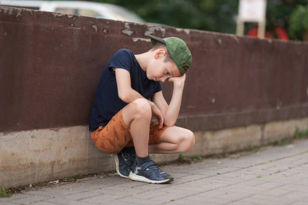 A teenage boy is squatting leaning against a concrete wall on the street. Upset and sad. Bullying. Quarrel with parents A teenage boy is squatting leaning against a concrete wall on the street. Upset and sad. Bullying. Quarrel with parents. sad teenager boy crying outdoors in park stock pictures, royalty-free photos & images