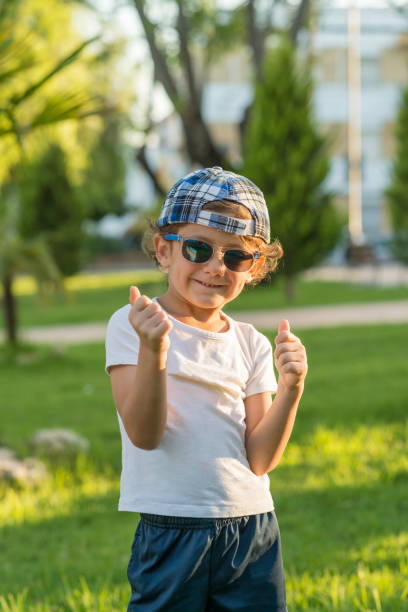 Portrait of a child posing in a park dressed in summer clothes, cap and sunglasses, making the thumbs up gesture.
