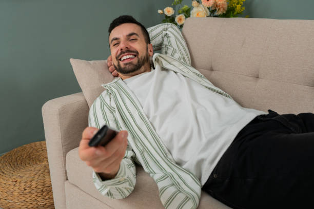 istock Smiling man relaxing on a sofa while watching a TV show, holding a remote control, enjoying leisure time in a cozy and modern living room at home 2200437866
