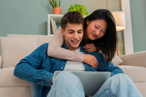 Friendly young wife embracing her husband while using laptop shopping online at web sitting on sofa at their cozy apartment. Shot of a couple in love looking at computer screen planning wedding
