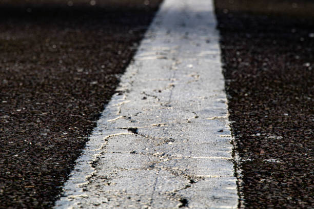 A strip of old white textured road markings. An old white road marking line runs along a dark asphalt surface. The paint is worn and textured, showing signs of age and weathering with visible cracks and chips. The rough texture of the asphalt contrasts with the faded white paint. This type of road marking is typically used for lane separation or traffic guidance. The image captures the close-up details of the road surface, highlighting the interplay between the materials and the effects of wear over time.