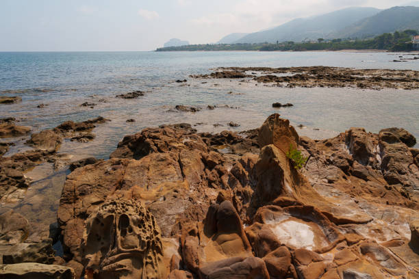 A coastal scene showcasing eroded rock formations near the water, with distant hills fading into the horizon. The rugged shoreline blends with the calm sea under a partly cloudy sky