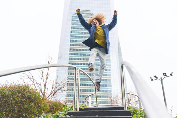 Joyful young man with curly hair jumping energetically on stairs in front of a modern skyscraper, exuding happiness and freedom