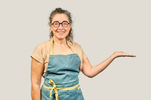 Friendly mature woman wearing apron and glasses is showing something on the palm of her hand, isolated on gray background