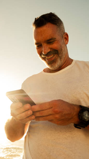 istock Smiling Man Using Smartphone at the Beach During Golden Hour 2200485851