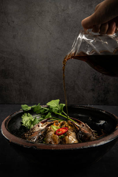 Pouring soy sauce into steamed fish in clay pot on black background