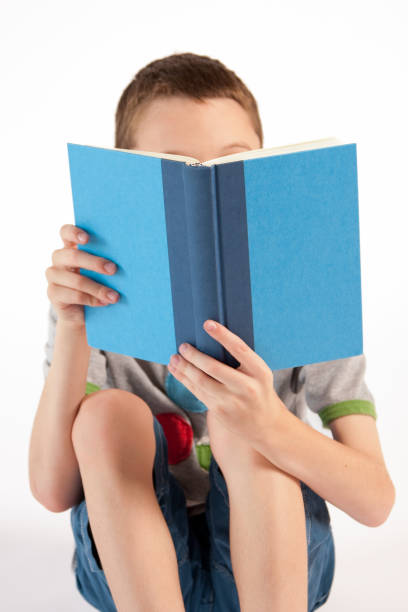 Young boy reading a book with face hidden behind the book. White background.