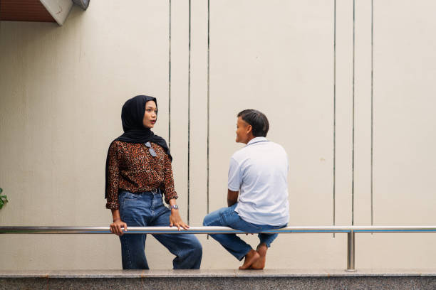A young Muslim woman wearing a stylish hijab and casual outfit engages in a friendly conversation with her friend, who is casually dressed in a white polo shirt and jeans. They are sitting on a railing in an outdoor urban setting, sharing a lighthearted moment. The relaxed body language and genuine smiles convey warmth, connection, and everyday friendship. The background features a minimalist wall.