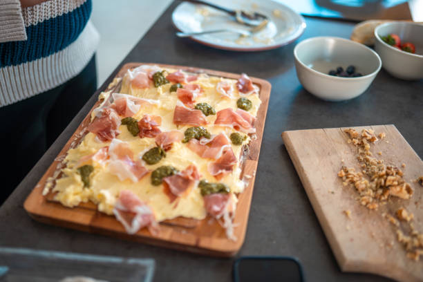 A butter board topped with creamy butter, pesto, and prosciutto is being prepared in a home kitchen. Nearby, bowls of fresh olives and tomatoes add to the vibrant spread.