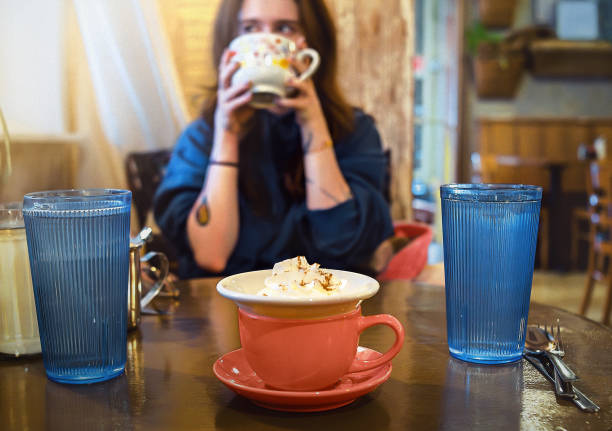 24-year-old woman drinks from a decorative cup at a coffee shop table. Two tall blue glasses and a red coffee cup with saucer sit on the wooden table surface. The cafe setting features wooden walls and casual dining atmosphere.