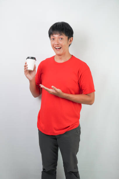 Excited young asian man wearing red shirt is holding a disposable coffee cup, isolated over white background.