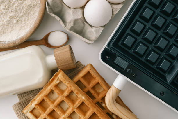 istock Ingredients for homemade waffles arranged on a white surface, including flour, sugar, eggs, and milk. A waffle maker with wooden handles is visible in the frame 2200522799