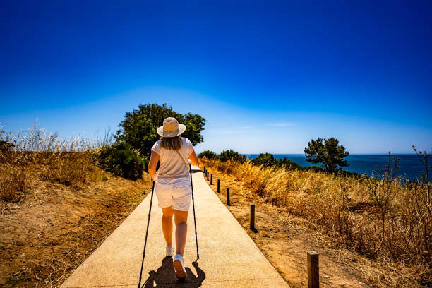 Summer vacation in Lagos Portugal. Mature woman practicing Nordic walking on path on cliffs during sightseeing Ponta da Piedade on beautiful sunny day. Back view