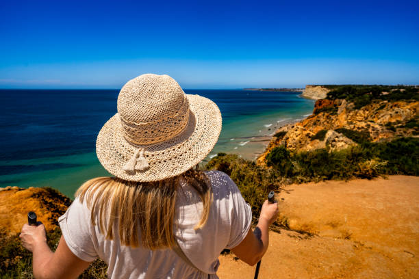 Summer vacation in Lagos Portugal. Mature woman practicing Nordic walking on path on cliffs during sightseeing Ponta da Piedade on beautiful sunny day. Back view.