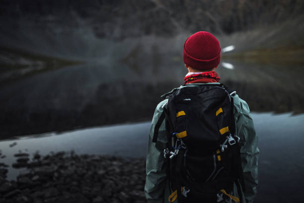 A traveler wearing a backpack observes the calm waters of a mountain lake at dusk. The serene environment frames the person against the reflective surface, emphasizing solitude and connection with nature.