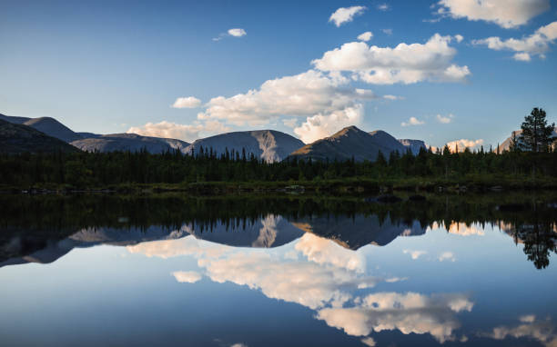 A calm lake reflecting lush forest, majestic mountains, and soft clouds on a sunny day. Nature's untouched beauty is showcased in this peaceful and picturesque landscape, offering a sense of tranquility and awe.