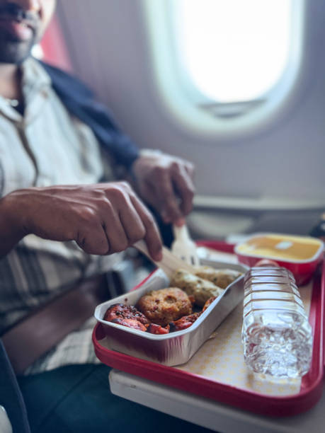 istock Close-up image of unrecognisable man in plane seat, fold down table containing plastic tray of chicken tikka, kofta (lamb meatball) and fried potato cake in disposable, single-use foil tray, bottle of water, cramped space, focus on foreground 2200568559