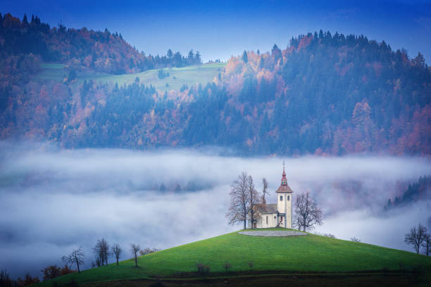 This dreamy morning view of Sveti Tomaz Church in Slovenia features mist-covered hills, golden autumn trees, a soft blue sky, rolling green meadows, and a peaceful, enchanting mountain landscape.