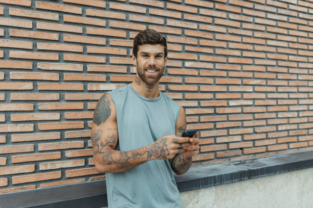A confident man with tattoos stands smiling against a plant backdrop, reflecting a cool demeanor and personal expression through his unique style and artistry.