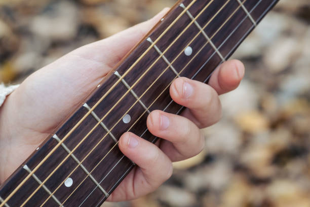 A close-up of a hand positioned on a guitar fretboard, illustrating finger placement. The person is holding down specific strings with fingertips, demonstrating a gripping technique. The guitar has six strings and visible frets, with tuning pegs partially visible. The hand is positioned near the guitar's headstock, indicating a chord or note is being played. The background is neutral, focusing attention on the hand and fretboard interaction. The strings are evenly spaced, and the fretboard is dark-colored.