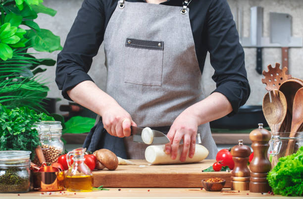 istock Home chef cuts big white radish. Cozy kitchen with wooden table, kitchenware, vegetables, herbs and ingredients for cooking. Healthy vegan eating, culinary, recipes, blogging 2200592427