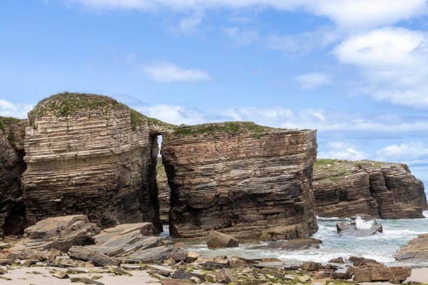 istock Impressive rock formations with layered sedimentary cliffs and a natural arch stand against the ocean. Waves crash on the rugged coastline under a blue sky 2200598855