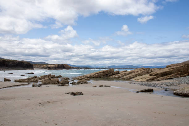 istock Scenic coastal landscape with sandy beach, unique rock formations, and ocean waves under a partly cloudy sky. Distant mountains enhance the natural beauty 2200598895