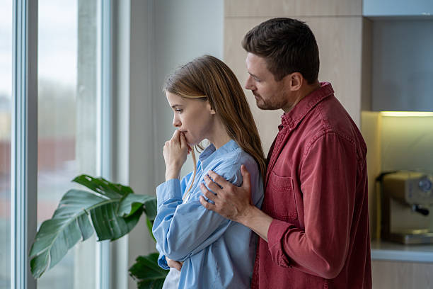supportive husband hugging his upset, depressive wife from behind at window in bright apartment - samimi olma stok fotoğraflar ve resimler