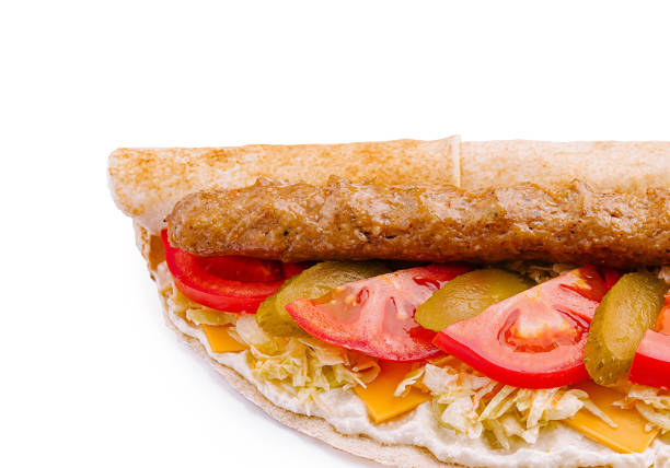 A hearty sandwich showcases a grilled sausage on top of melted cheese, layered with fresh tomato slices, crunchy pickles, and shredded lettuce, served on a flatbread.
