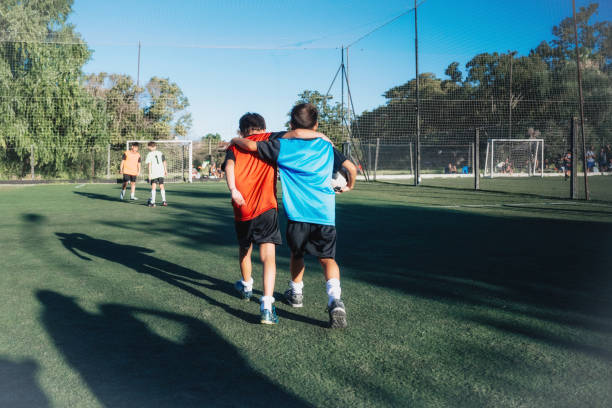 rear view on two boys from opponent teams walking embraced after match on pitch