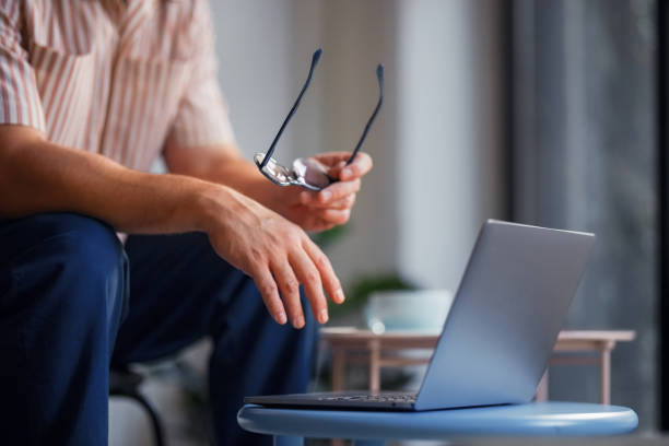 Focused Individual Holding Glasses While Looking at a Laptop on a Coffee Table