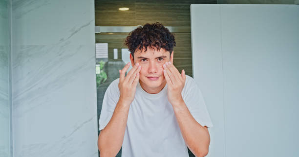 istock Young man smiling while applying moisturizer to his face in front of mirror in the bathroom at home. Healthy lifestyle in morning routine concept. 2200648078